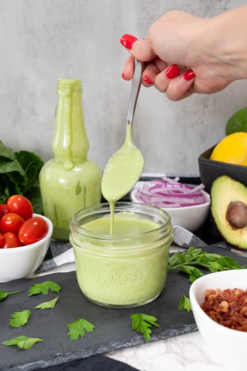 A spoon drizzles creamy green goddess dressing from a mason jar, surrounded by a bottle of the same dressing, sliced red onions, halved cherry tomatoes, a halved avocado, parsley, and a bowl of bacon bits, all arranged on a dark slate board.