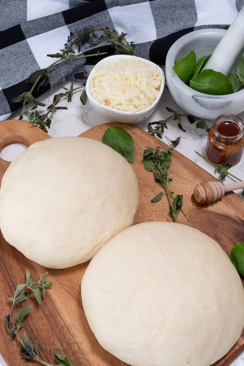 Two balls of pizza dough rest on a wooden cutting board with fresh herbs scattered around. Nearby, there is a bowl of shredded cheese, a mortar and pestle with basil leaves, a small jar of honey with a wooden dipper, and a checkered napkin—perfect ingredients for an irresistible pizza dough recipe with honey.