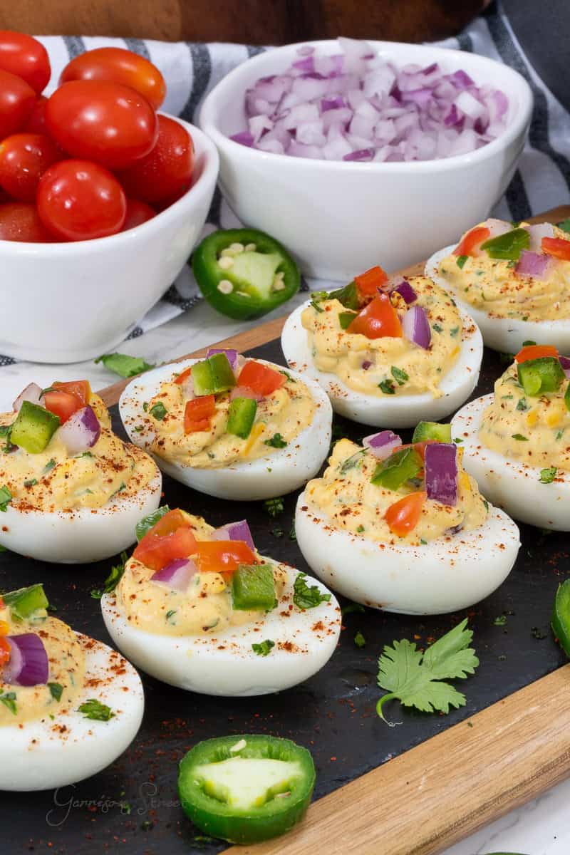 A platter of Mexican taco deviled eggs garnished with chopped jalapeños, red onions, and tomatoes. The eggs are sprinkled with paprika and fresh herbs. In the background, there are bowls containing chopped red onions and cherry tomatoes, alongside whole jalapeño slices.