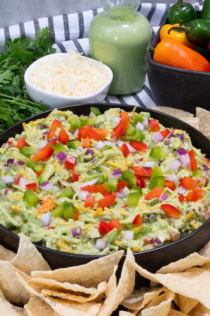 A close-up of a black bowl filled with layered green goddess dip topped with diced red and green peppers, and red onions. Surrounding the bowl are tortilla chips, a bowl of shredded cheese, a jar of green goddess dressing, fresh parsley, and colorful bell peppers.