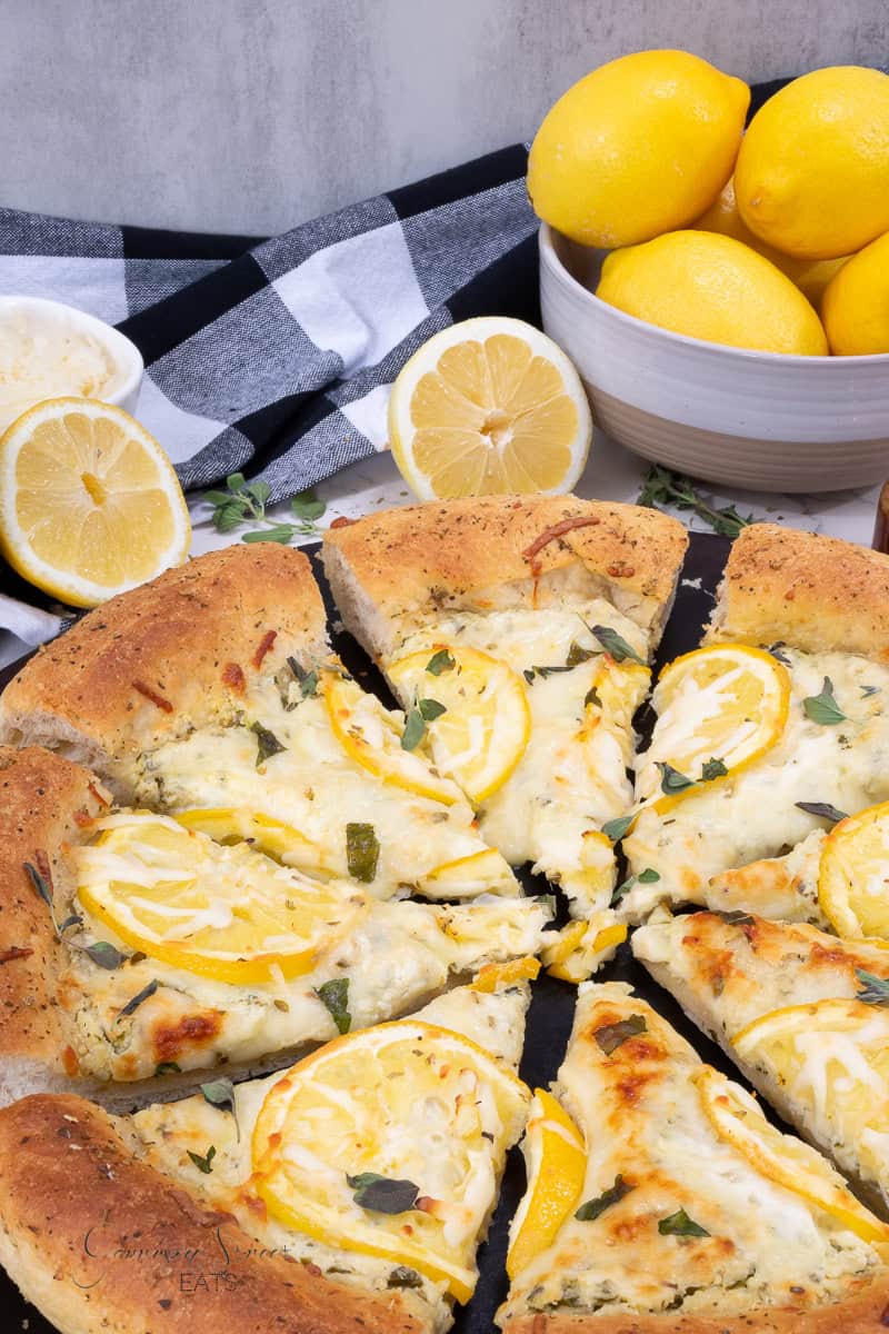 A freshly baked Lemon Ricotta Pizza rests on a dark surface, topped with lemon slices, herbs, and cheese. A black and white checkered cloth, a bowl filled with bright yellow lemons, and a sliced lemon are visible in the background, along with a small dish of grated cheese.