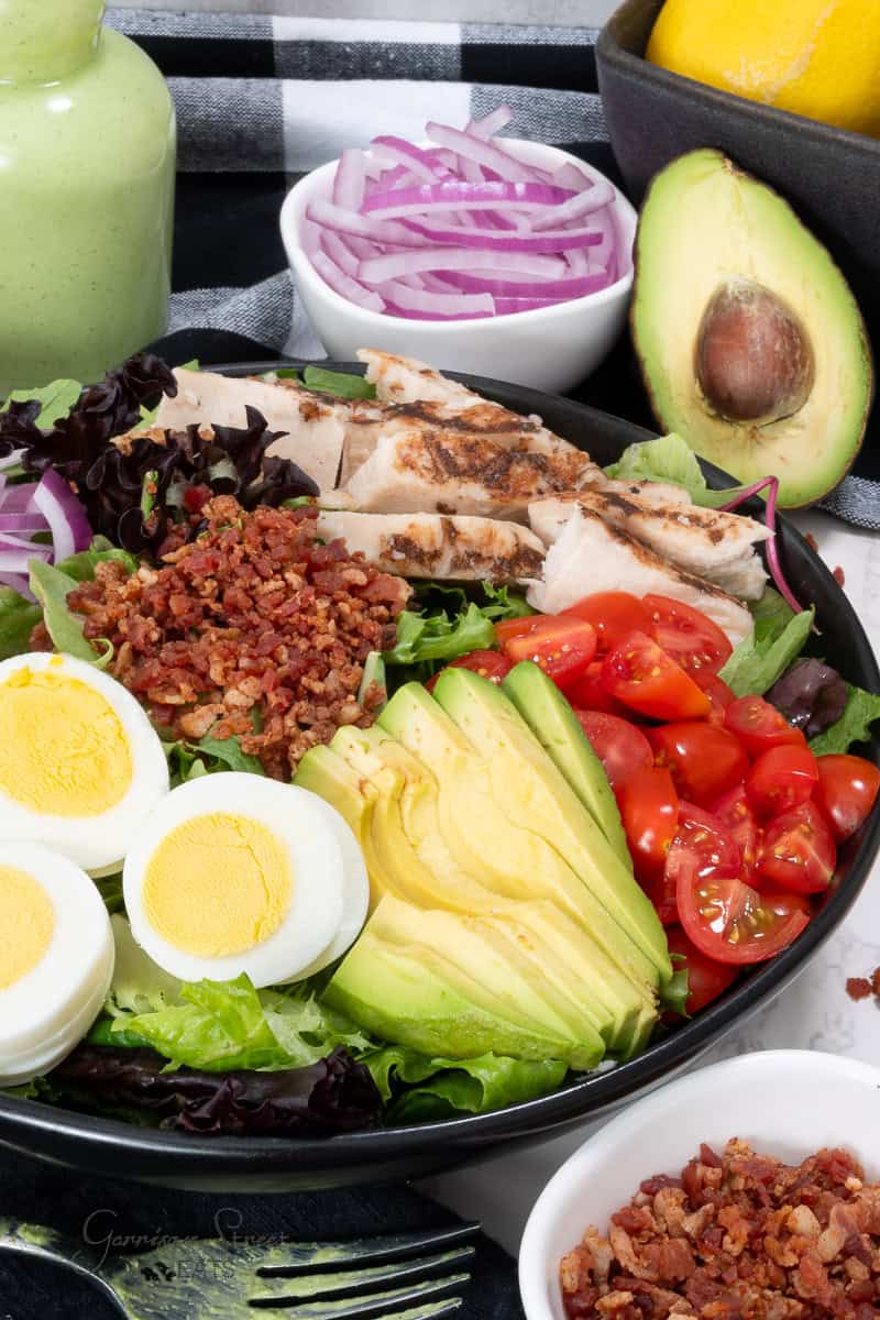 A close-up of a colorful Cobb salad featuring grilled chicken slices, halved cherry tomatoes, avocado slices, hard-boiled egg halves, crumbled bacon, and mixed greens. Surrounding the salad are ingredients including red onion slices, a whole avocado, and lemons; inspired by the Panera Green Goddess Salad recipe.