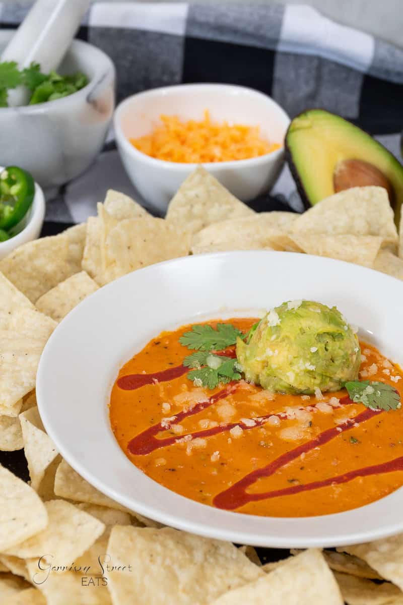 A bowl of creamy cheese dip topped with a scoop of guacamole, cilantro, and hot sauce drizzle, surrounded by tortilla chips. Reminiscent of a Torchy Tacos queso recipe, the scene features bowls of shredded cheese, sliced jalapeños, and a half avocado in the background.