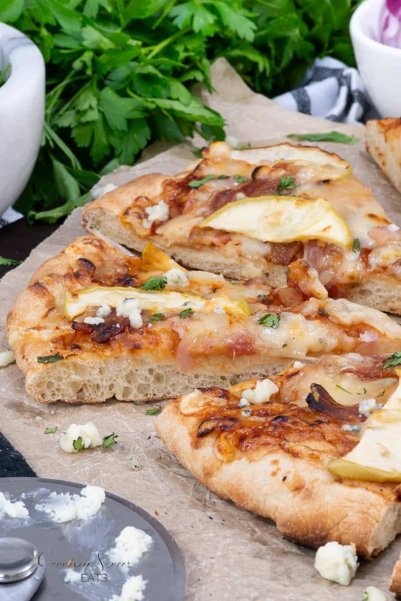 A close-up of a sliced pizza with a thick Poolish Pizza Dough crust with air bubbles with toppings featuring melted cheese, chunks of apple, and bits of bacon. The pizza rests on parchment paper and is surrounded by fresh parsley and a bowl, with a pizza wheel containing crumbled cheese in the foreground.