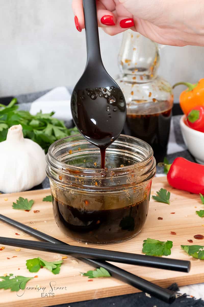 A hand with red painted nails holds a black spoon dripping with dark teriyaki sauce over a jar on a wooden cutting board. Surrounding the jar are fresh parsley leaves, a white garlic bulb, a red bell pepper.