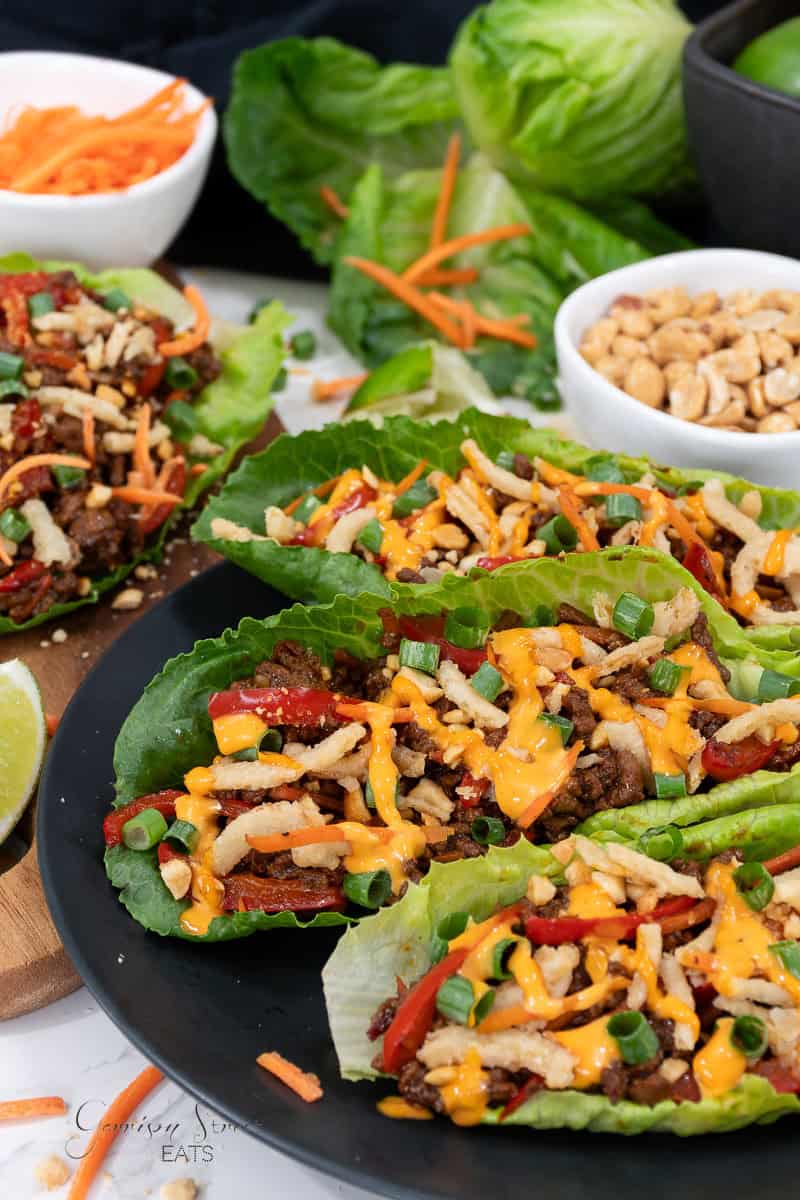 A plate of Asian lettuce wraps filled with seasoned ground beef, shredded carrots, red bell peppers, green onions, and crispy fried onions. Drizzled with sauce and placed on fresh romaine leaves. Additional ingredients sit in the background.