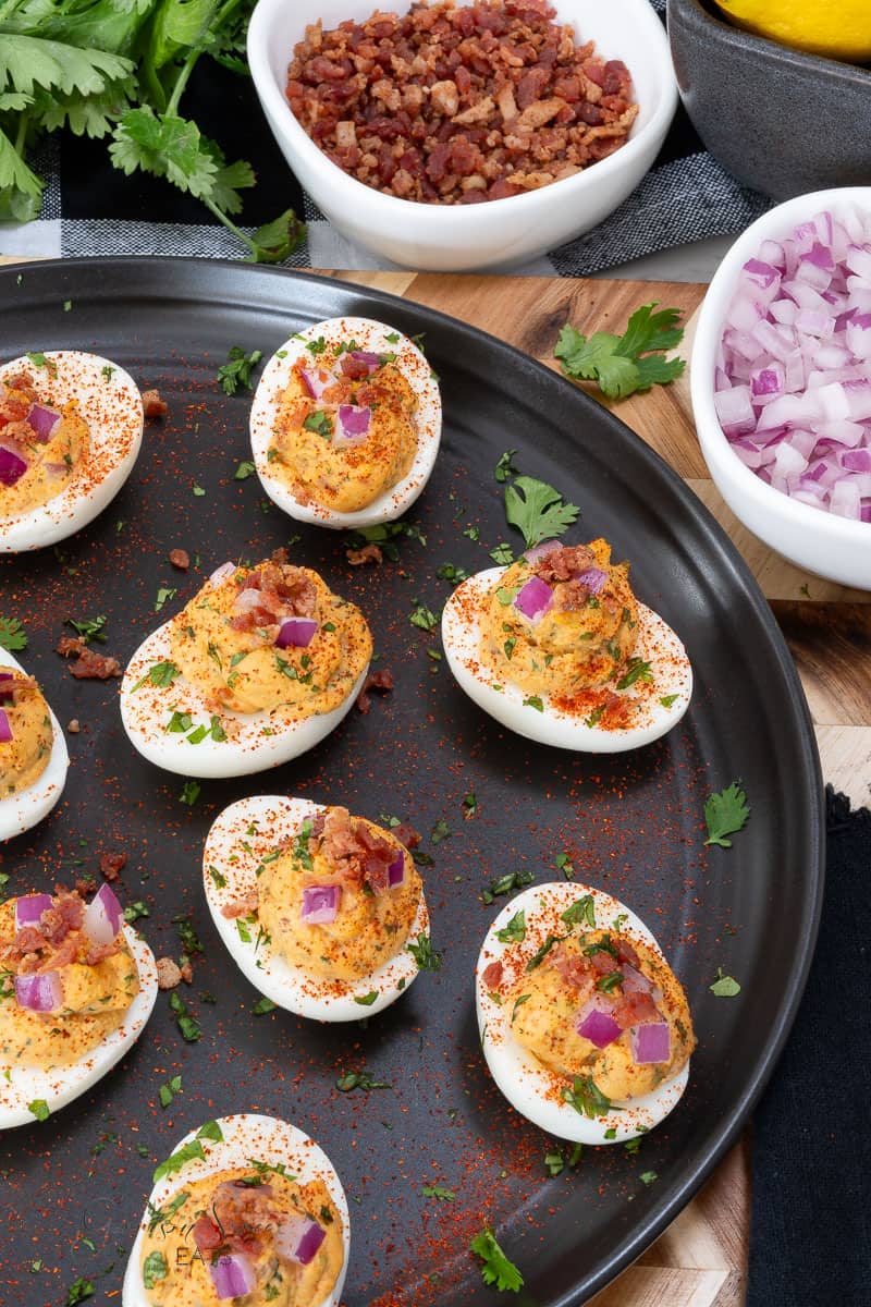 A black plate holds several BBQ deviled eggs garnished with paprika, red onion, and bacon bits on a wooden table. To the side, there are small bowls containing bacon bits and chopped red onions. Fresh cilantro leaves are scattered around the plate.