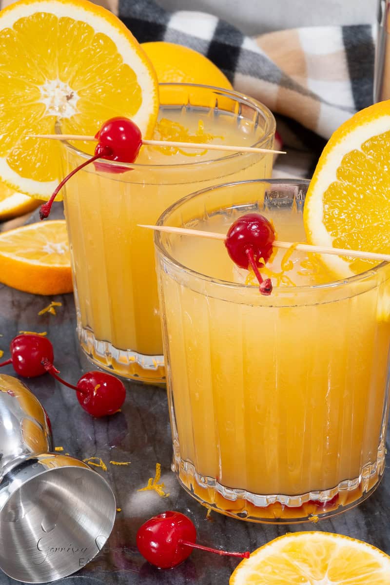 Three glasses of an orange-colored amaretto stone sour cocktail are garnished with orange slices and maraschino cherries on toothpicks. Fresh orange halves, cherries, and a stainless steel jigger are placed around the drinks on the table. A checkered cloth is slightly visible in the background.