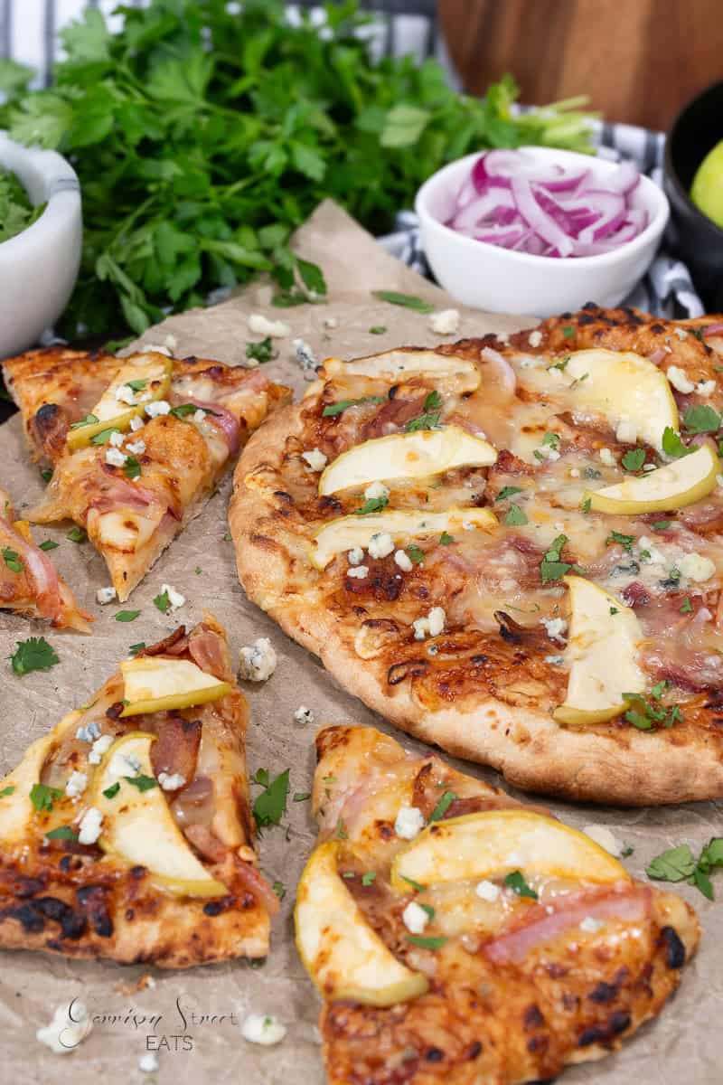 A blue cheese pizza topped with apple slices, cheese, and herbs is on a wooden surface. Two slices have been cut and slightly separated from the main pizza. In the background, there are bowls containing chopped onions and fresh greens. The wood is partly covered in parchment paper.