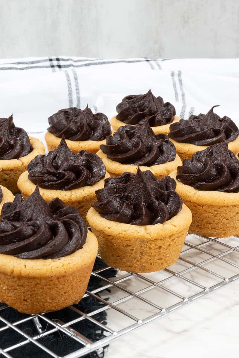 A close-up of a batch of cookie cups topped with rich chocolate frosting, made from an easy chocolate icing recipe, arranged on a cooling rack. The cookie cups have a golden-brown exterior contrasting with the dark, creamy cocoa powder frosting. A checkered cloth is partially visible in the background.