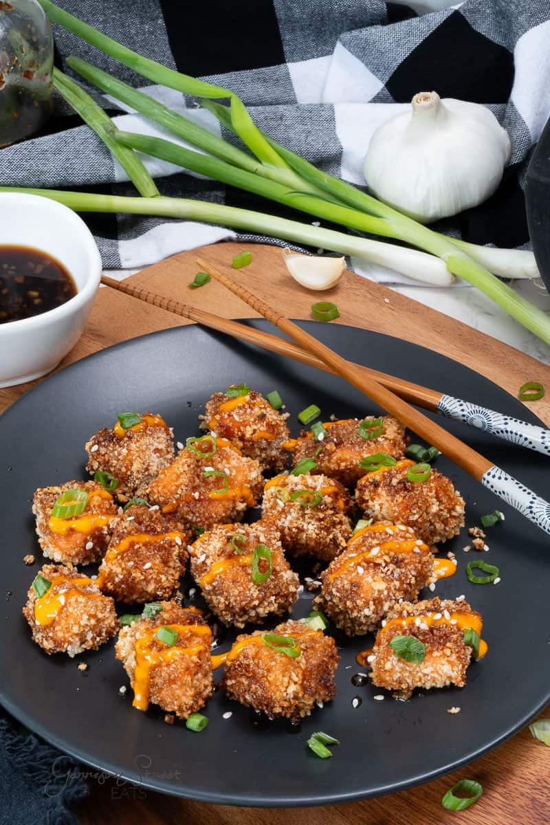 A black plate of crispy fried salmon bites garnished with green onions and drizzled with an orange sauce. Chopsticks rest on the side of the plate. In the background, there's a bowl of dipping sauce, a whole garlic bulb, green onions, a napkin, and a wooden board—perfect for your next Ninja air fryer salmon recipe.