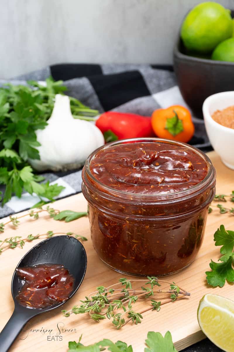 A jar of homemade Caribbean jerk sauce sits on a wooden cutting board. A spoon with a dollop of the piquant sauce is next to the jar. Surrounding the items are fresh herbs, garlic, mini sweet peppers, lime, and a small bowl of seasoning, all set against a checkered cloth background.