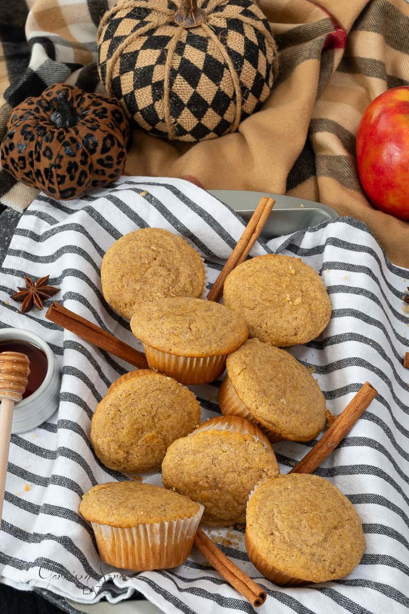 A white dish towel with black stripes holds freshly baked Pumpkin Spice Cornbread muffins, arranged with cinnamon sticks. An apple, two fabric pumpkins with checkered and leopard prints, a honey dipper, and a small bowl of honey are placed around the muffins as part of the autumn-themed setup.