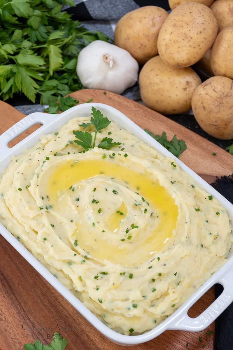 A dish of the best garlic herb mashed potatoes, topped with a swirl of melted butter, garnished with chives and a parsley sprig. In the background are whole potatoes, a bulb of garlic, and a bunch of fresh parsley, all on a wooden cutting board and gray cloth.