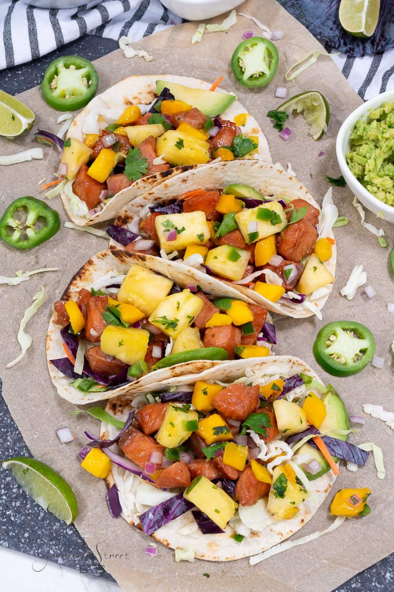 Three soft tacos filled with a colorful mix of diced mango, pineapple, red onions, bell peppers, shredded cabbage, and cilantro, placed on parchment paper. The Salmon Fish Tacos are garnished with slices of jalapeño and lime wedges, with a small bowl of guacamole on the side.