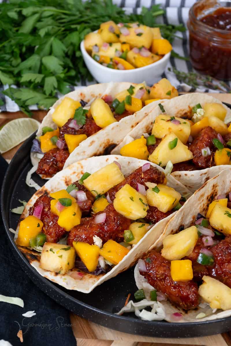 A plate of four tacos filled with crispy breaded chicken, diced mango, pineapple, red onions, jalapenos, and green herbs. A lime wedge, a bowl of mango salsa, and fresh cilantro are in the background. The tacos are served on a dark circular plate.