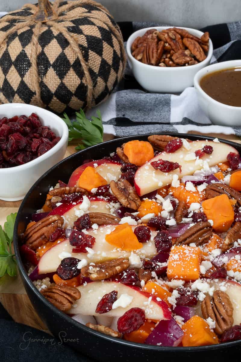 A fall quinoa bowl featuring roasted sweet potatoes, pear slices, pecans, dried cranberries, and feta cheese crumbles sits in a pan. Surrounding the dish are small bowls with extra dried cranberries, pecans, and dressing. A black and tan checkered pumpkin and greenery complete the autumnal setting.