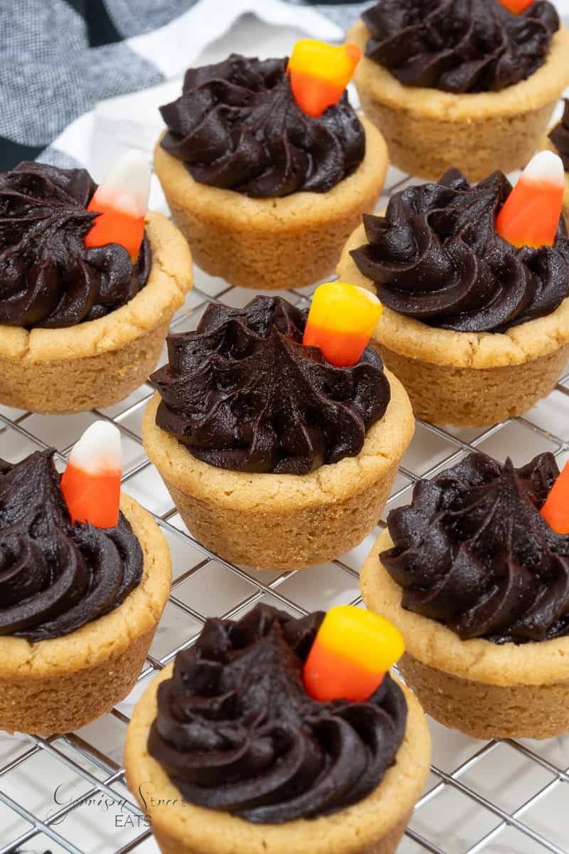 A cooling rack of Halloween themed cookie cupcakes is shown. The cupcakes have a cookie base, topped with swirls of dark chocolate frosting and decorated with candy corn pieces. The background features a black and white patterned cloth.