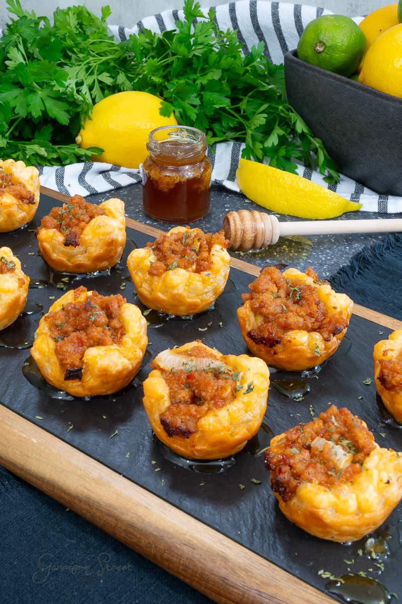 A slate board displays savory puff pastry tarts filled with a golden mixture of sausage, onion, and goat cheese, topped with honey. Fresh parsley, bright yellow lemons, limes, and a small jar of honey are in the background, with a honey dipper placed nearby.