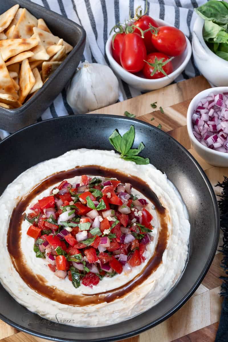 A black bowl filled with creamy dip topped with diced tomatoes, onions, and herbs. Surrounding the bowl are diced onions, cherry tomatoes, pita chips, basil leaves, and a garlic bulb on a striped cloth backdrop.