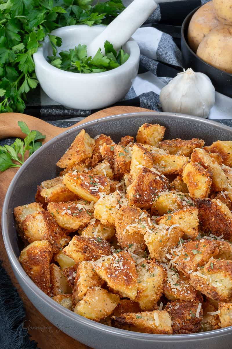 A bowl of crispy, golden roasted parmesan potatoes garnished with grated cheese and herbs. Fresh parsley, garlic, and whole potatoes are in the background, along with a mortar and pestle.