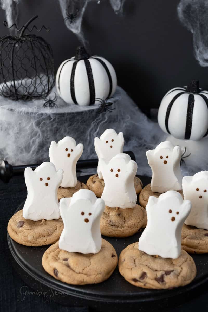 A spooky Halloween scene with marshmallow ghosts on chocolate chip snickerdoodle cookies. Black and white pumpkins and spider web decor are in the background, creating a festive atmosphere.