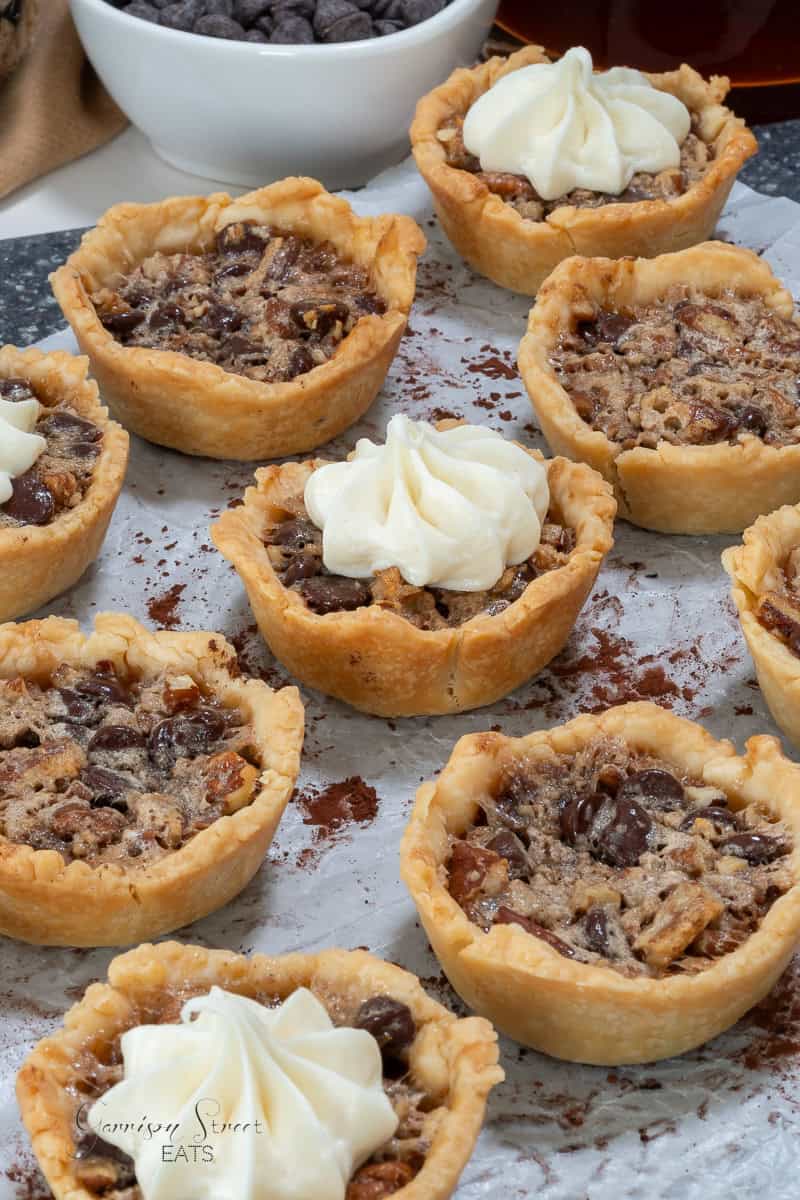 A selection of small, baked pies filled with pecans and chocolate chips, topped with whipped cream. The pies are arranged on a speckled surface.