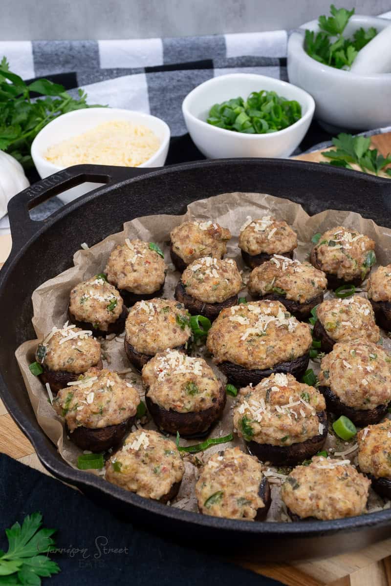 A skillet filled with stuffed mushrooms topped with parmesan cheese and herbs. Green onions and grated cheese in small bowls are nearby, along with fresh parsley. A black and white checkered cloth is in the background.