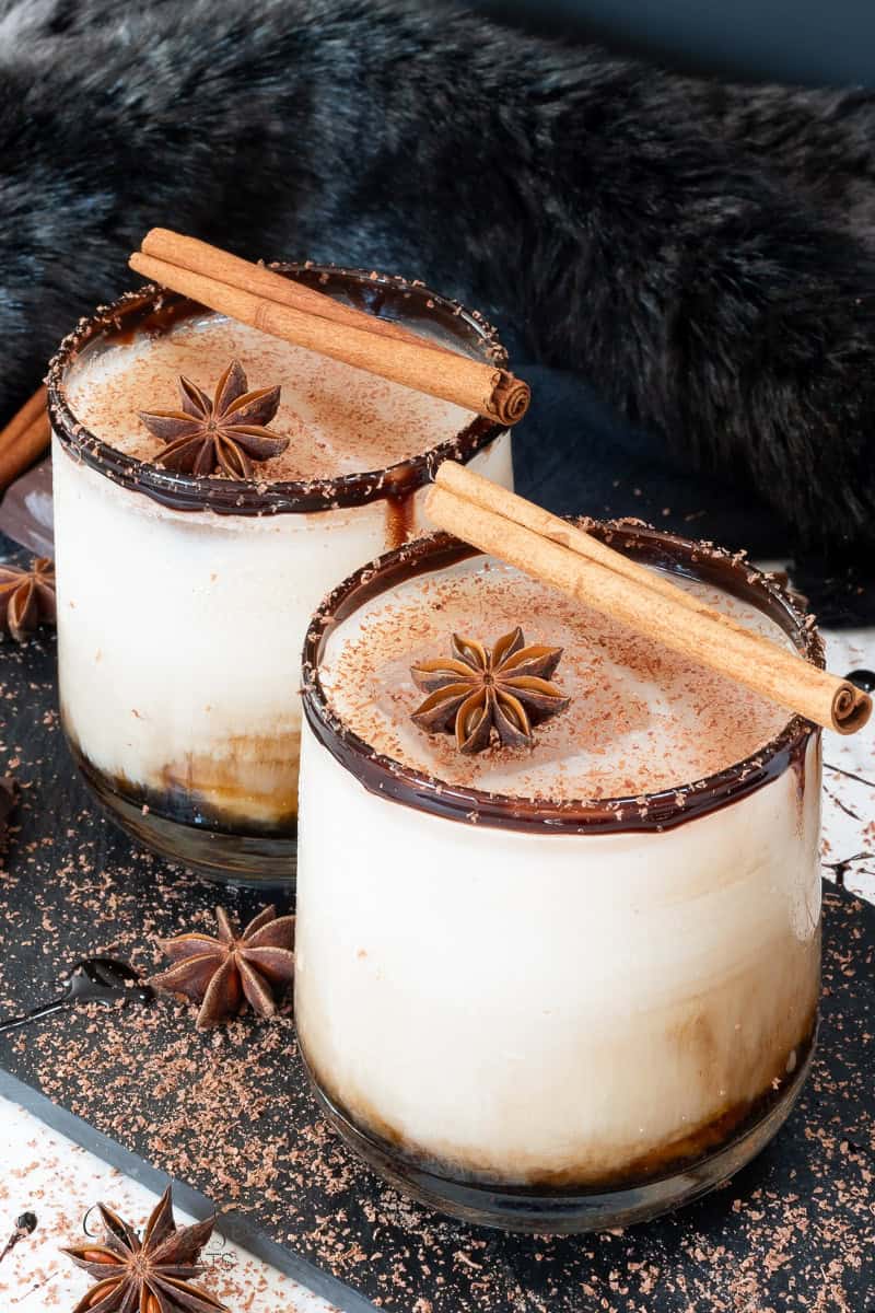 Two glasses of creamy white Russians topped with cinnamon sticks and star anise sit on a dark surface. The rims are coated with chocolate sauce and shavings, and a fluffy black background enhances the cozy feel. Star anise and grated chocolate surround the glasses.