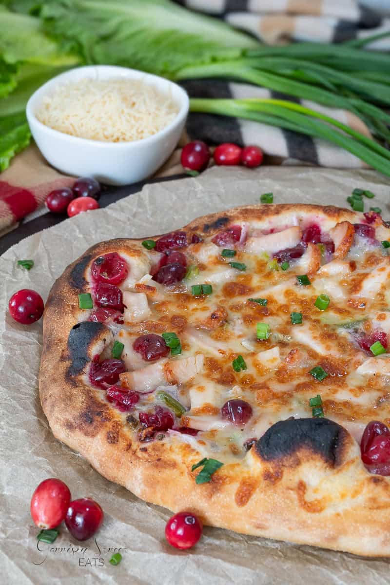 A freshly baked pizza topped with melted cheese, cranberries, and green onions on a parchment-lined surface. Fresh cranberries and a small bowl of grated cheese are in the background.