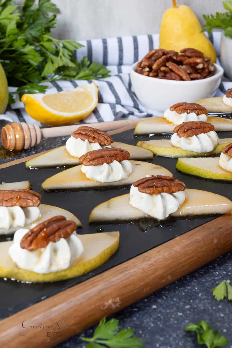 Slices of pear topped with dollops of whipped blue cheese and pecans are arranged on a slate board. A lemon wedge and bowl of pecans are in the background, with greenery surrounding the scene.