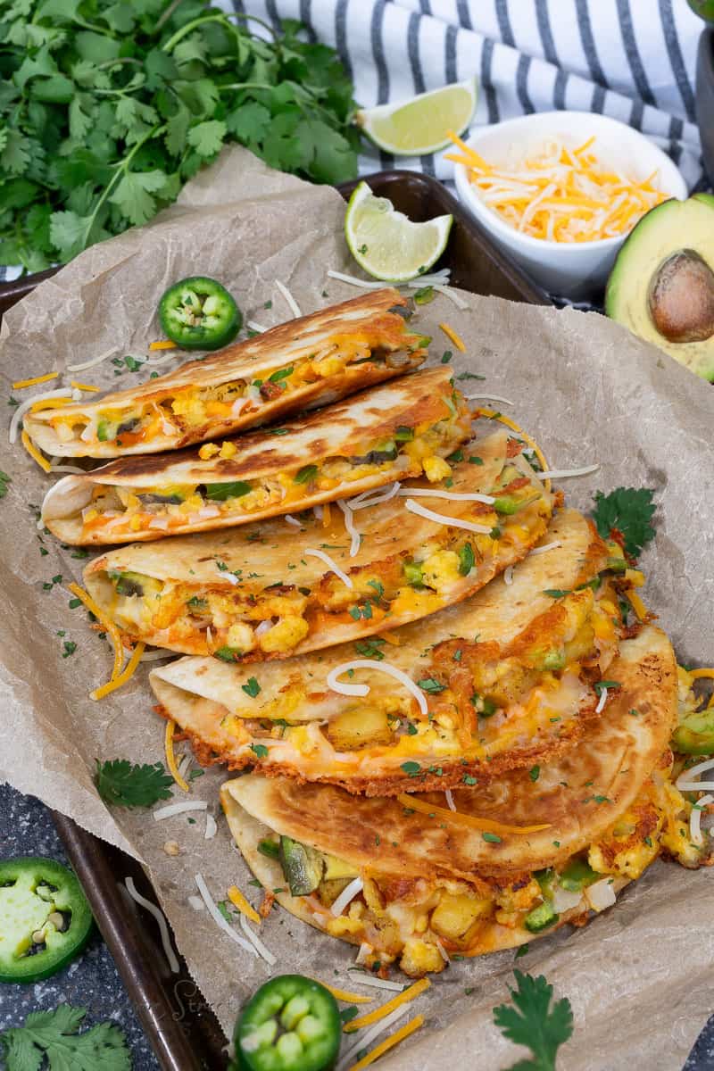A tray of crispy, golden-brown tacos filled with cheese, potatoes, and eggs. Garnished with sliced jalapeños and cilantro, accompanied by a halved avocado, lime wedges, and a small bowl of shredded cheese in the background.