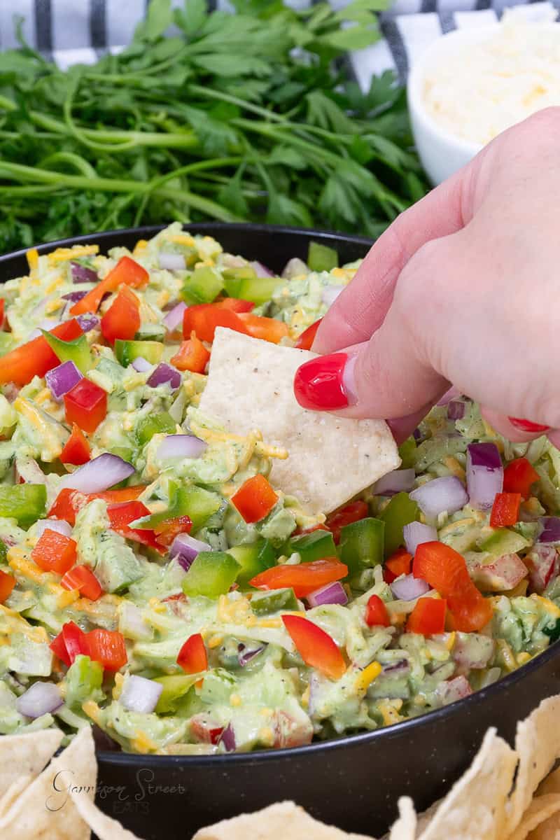 A hand with red-painted nails dips a tortilla chip into a colorful green cheese dip topped with chopped red onions, green bell peppers, red bell peppers, and shredded cheese. Fresh parsley and a bowl of dip are in the background.