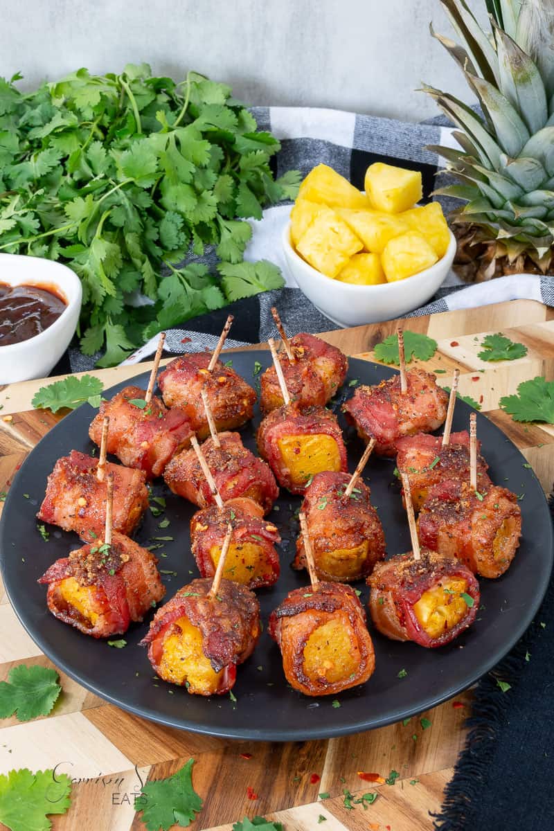 A black plate filled with bacon wrapped pineapple bites, each held with a toothpick. The plate is garnished with herbs and placed on a wooden board. In the background are fresh cilantro, a bowl of pineapple chunks, and a bowl of dark sauce.