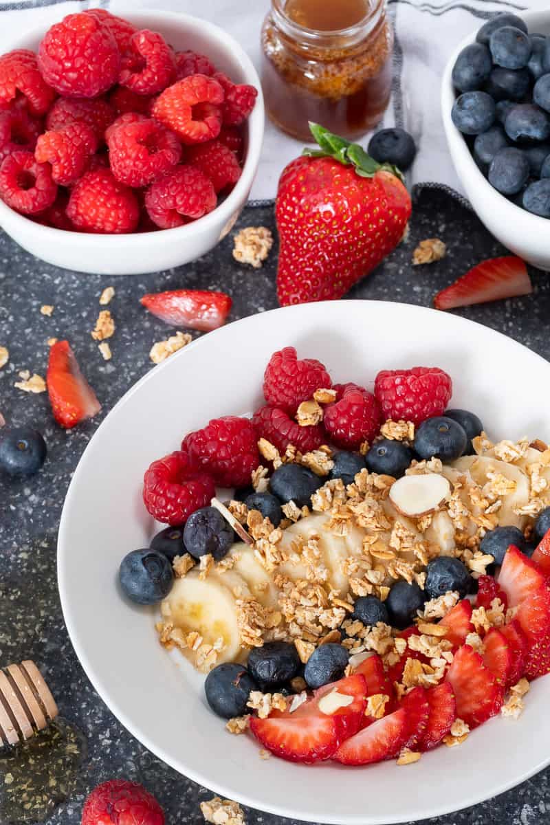 A bowl of Greek yogurt topped with granola, sliced bananas, strawberries, blueberries, and raspberries. Surrounding the bowl are dishes of fresh raspberries and blueberries, a whole strawberry, and a jar of honey with a drizzle stick.