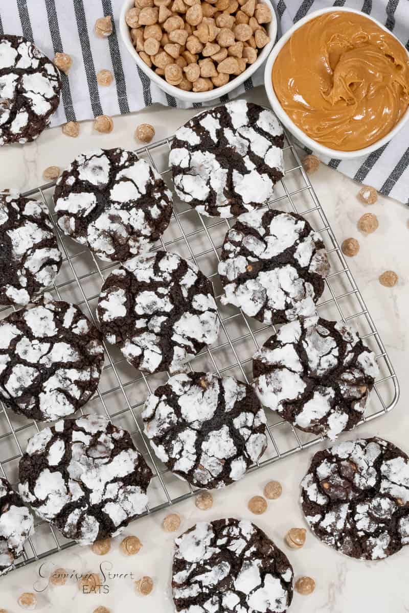 Chocolate crinkle cookies with a powdered sugar coating are cooling on a wire rack. Nearby are a bowl of peanut butter chips and a bowl of peanut butter. The setup is on a light-colored marble surface with a striped cloth.