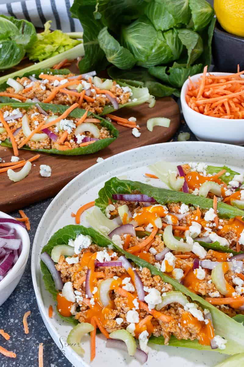 Fresh lettuce wraps filled with ground buffalo chicken, shredded carrots, sliced red onions, and crumbled blue cheese. Drizzled with orange sauce, the wraps are surrounded by bowls of more ingredients. A vibrant and healthy meal presentation.