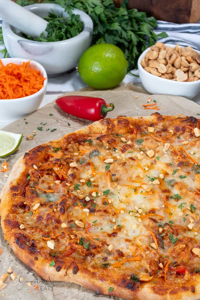 A kung pao chicken pizza topped with melted cheese, shredded carrots, and chopped peanuts is on parchment paper. Surrounding it are bowls of carrots, peanuts, a mortar with pestle, herbs, a lime, and a red chili pepper.