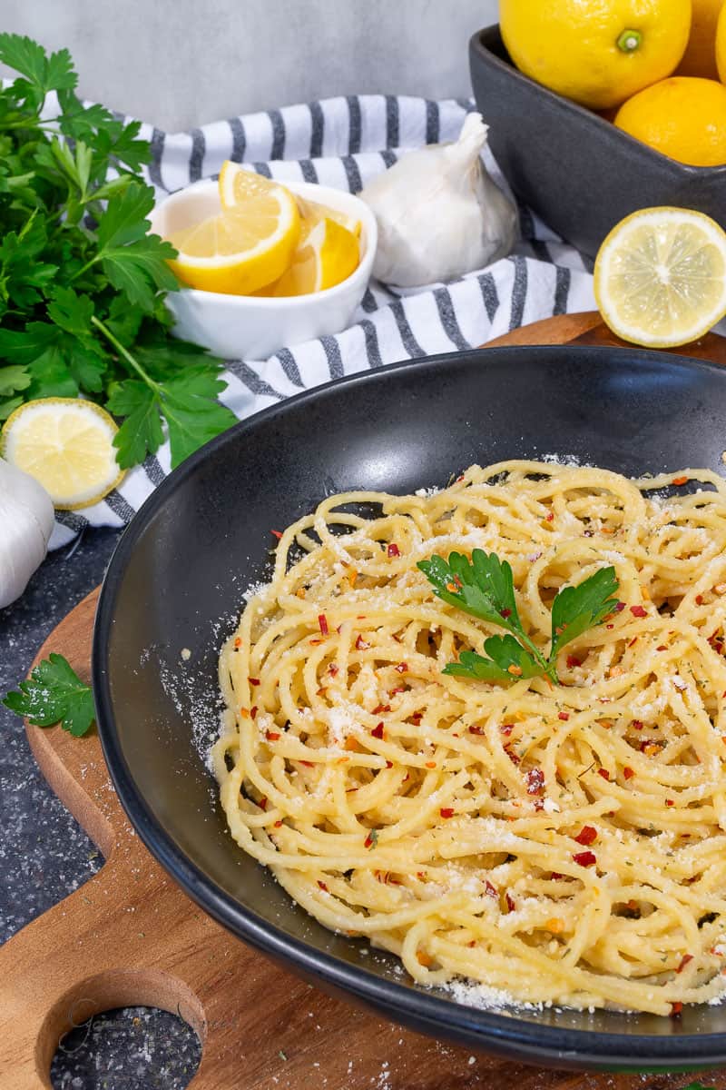 A black bowl filled with spaghetti parmesan cheese buttered noodles, topped with red chili flakes and a parsley garnish. In the background, there are lemons, garlic, and parsley on a striped cloth, and a wooden board beneath the bowl.