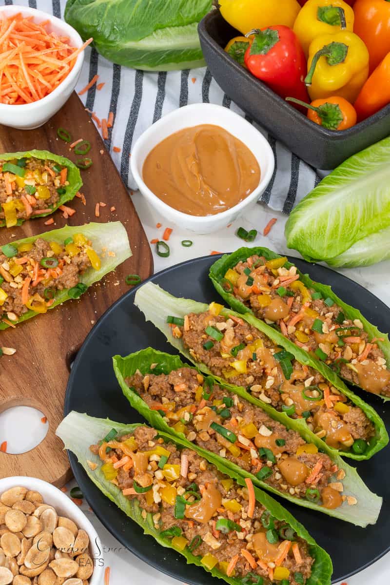 Thai peanut lettuce wraps filled with ground turkey, chopped vegetables, and peanuts are served on a black plate. A cutting board displays additional wraps and shredded carrots. A bowl of peanut sauce and a bowl of mini bell peppers are in the background.