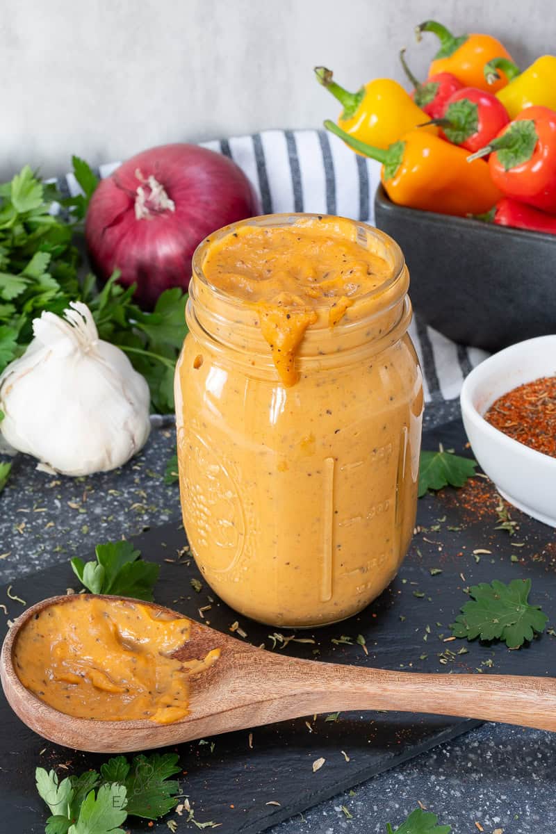 A jar of orange creamy Cajun alfredo sauce is placed on a slate, surrounded by fresh garlic, parsley, an onion, and colorful bell peppers. A wooden spoon with sauce rests beside the jar, and a bowl of seasoning is nearby.