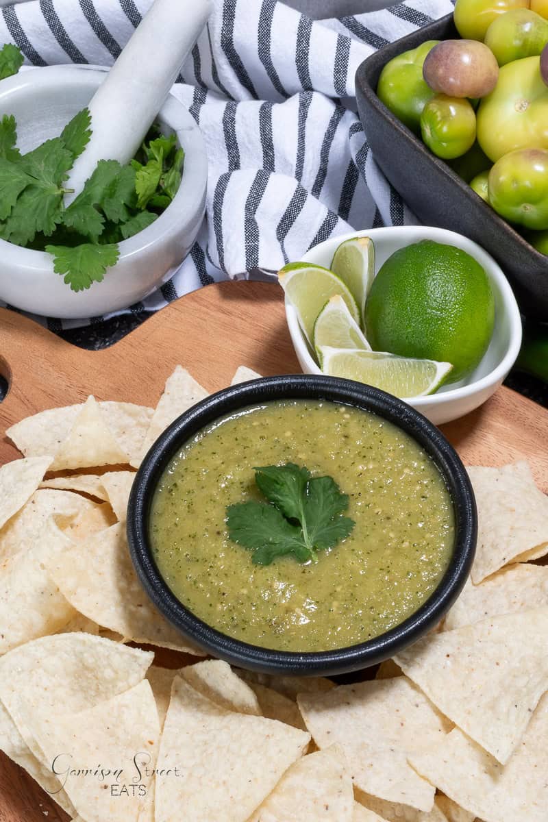 A bowl of green tomatillo salsa garnished with cilantro sits surrounded by tortilla chips. Nearby are lime wedges, a whole lime, fresh tomatillos, and a mortar with cilantro, all on a wooden board with a striped cloth.