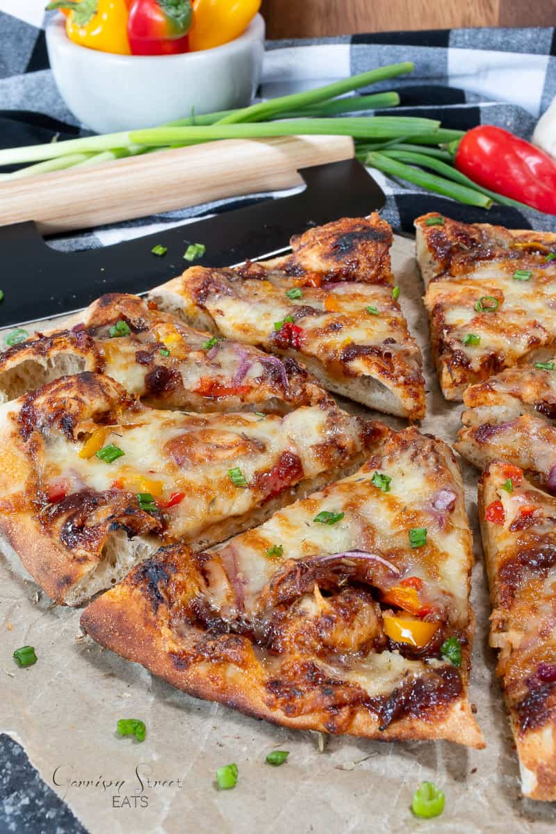 A close-up of a sliced homemade pizza topped with melted cheese, red onions, and colorful bell peppers. The pizza is on parchment paper, with green onions and fresh vegetables in the background.