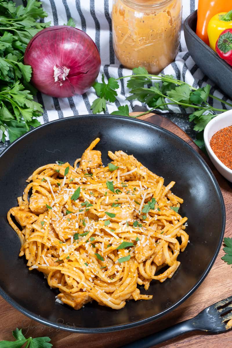 A black bowl filled with creamy pasta topped with grated cheese and chopped parsley sits on a wooden surface, surrounded by fresh parsley, a red onion, a jar of sauce, and spices.