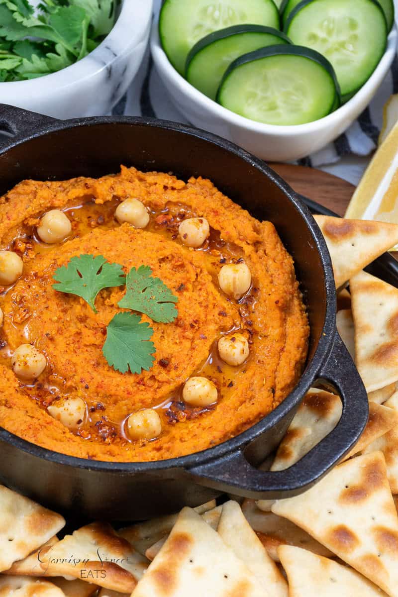 A cast iron skillet filled with roasted red pepper hummus, garnished with chickpeas, olive oil, chili flakes, and cilantro. Surrounded by pita bread chips, cucumber rounds, and a bowl of fresh cilantro.