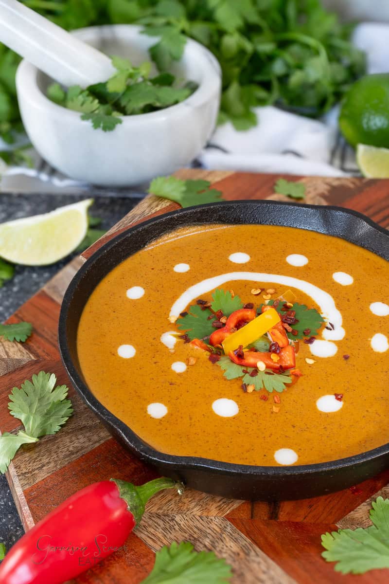 A cast iron pan of creamy orange curry sauce garnished with white sauce dots, cilantro, sliced red and yellow chili peppers, and red pepper flakes, placed on a wooden board with fresh herbs and lime in the background.