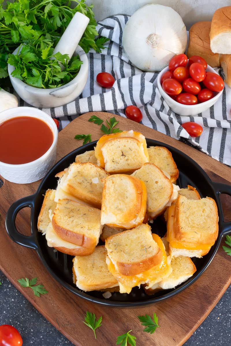 A black plate filled with mini grilled cheese sandwiches sits on a wooden board, surrounded by a small cup of tomato soup, cherry tomatoes, garlic, fresh parsley, and a striped kitchen towel.