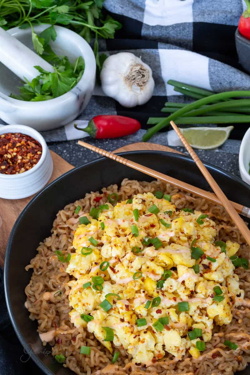 A bowl of brothless ramen recipe topped with scrambled eggs, chopped green onions, and red pepper flakes, with chopsticks resting on the bowl. Fresh herbs, garlic, chili peppers, and spices are visible in the background.