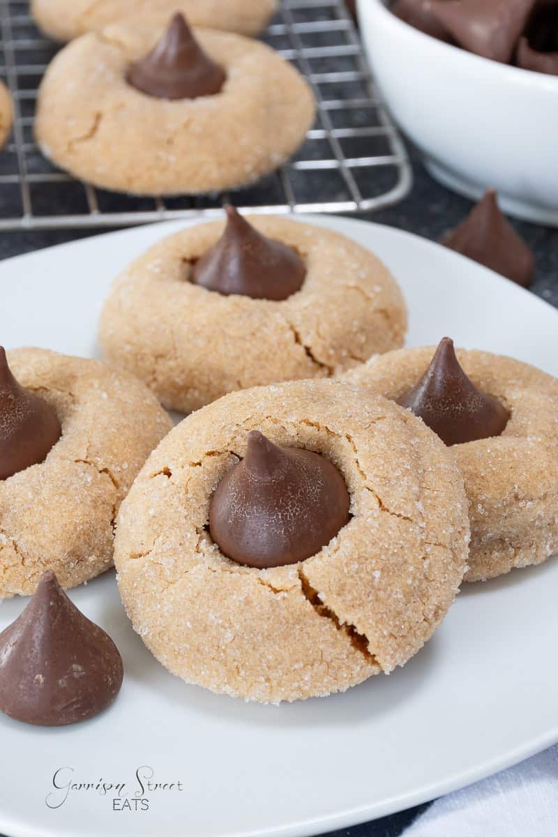 A plate of peanut butter blossom cookies, each topped with a chocolate kiss, sits in the foreground. More cookies and a bowl of chocolate kisses are visible in the background.