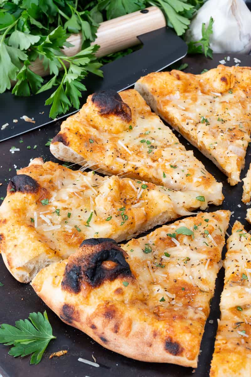 Close-up of a sliced cheesy Cajun chicken pizza garlic pizza topped with herbs on a dark surface. Fresh parsley, a garlic bulb, and a grater are visible in the background.
